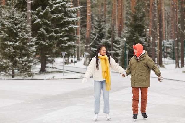 Felice coppia multietnica pattinaggio sulla pista di pattinaggio nella foresta invernale durante le vacanze invernali