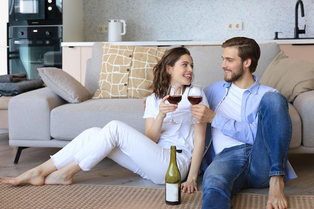 Felice coppia seduta rilassante sul pavimento in soggiorno a bere vino rosso Sorridente giovane marito e moglie riposano a casa godersi insieme un appuntamento romantico durante il fine settimana in famiglia