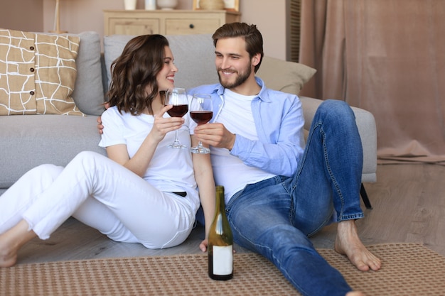 Felice coppia seduta, rilassante sul pavimento in soggiorno, bevendo vino rosso. Il giovane marito e la moglie sorridenti riposano a casa e godono insieme di un appuntamento romantico durante il fine settimana in famiglia.