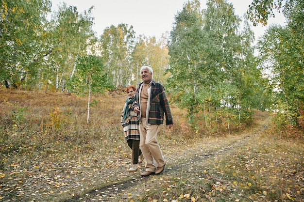 Felice coppia senior in abbigliamento casual caldo che si muove lungo la strada o il sentiero nel bosco mentre si gode una passeggiata nel fine settimana autunnale