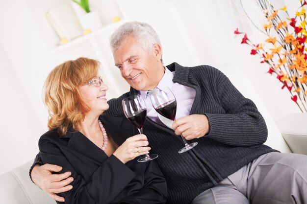 Felice coppia senior seduta abbracciata a casa, sorridente e bere vino rosso.