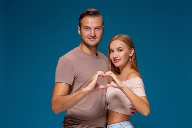 Felice coppia vestita in abiti casual, facendo a forma di cuore dalle dita, ritratto in studio su sfondo blu