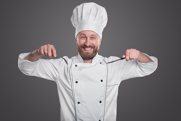 Felice cuoco maschio barbuto in uniforme bianca da chef e cappello che tiene forchetta e coltello mentre si prepara ad assaggiare il cibo su sfondo grigio