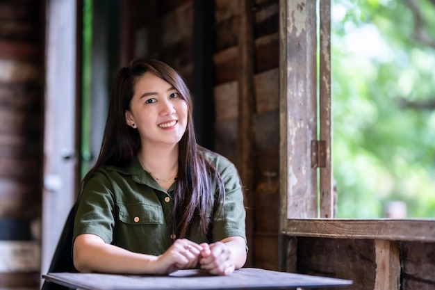 Felice del ritratto Felice bella donna asiatica sorridente che indossa un abito verde brunastro elegante hipster con accanto alla finestra di uno sfondo di casa in legno