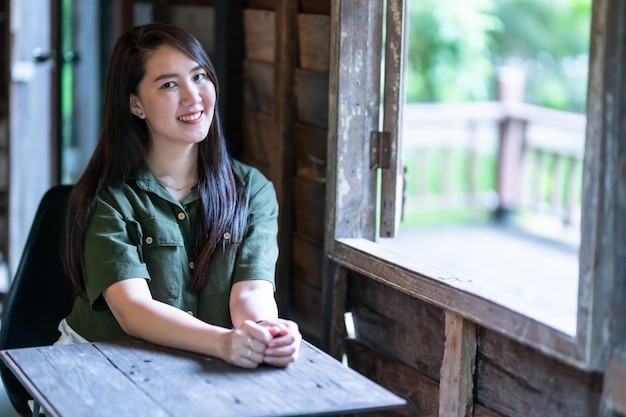 Felice del ritratto Felice bella donna asiatica sorridente che indossa un abito verde brunastro elegante hipster con accanto alla finestra di uno sfondo di casa in legno