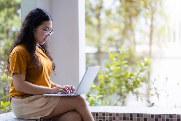 Felice di persone asiatiche freelance business casual femminile che lavora con il computer portatile seduto su una panchina in una caffetteria come lo sfondo