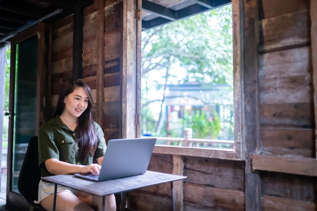 Felice di persone asiatiche freelance donna d'affari che indossa un abito verde brunastro elegante hipster casual che lavora con il computer portatile con una tazza di caffè accanto alla finestra di uno sfondo di casa in legno