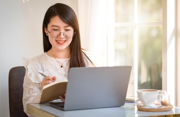 Felice di persone freelance asiatiche Donna d'affari che prende appunti scritti su un notebook che lavora casualmente con un computer portatile con una tazza da caffè e uno smartphone al bar, Stile di vita aziendale