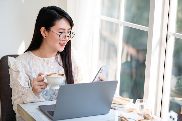 Felice di persone freelance asiatiche Donna d'affari che scrive un messaggio sullo smartphone e tiene in mano una tazza di caffè che lavora casualmente con il computer portatile al bar, Stile di vita aziendale