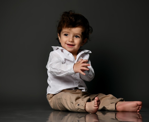 Felice divertente flirtare elegante bambino a piedi nudi che indossa pantaloni camicia che mostra la lingua Ritratto isolato girato su sfondo grigio studio Infanzia felice e moda Crescita e sviluppo del bambino