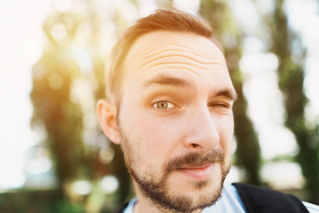 Felice divertente studente barbuto uomo in maglietta blu con zaino in piedi davanti a alberi verdi Allegro Hipster in posa per una foto sorridente guardando la telecamera rovinare un occhio