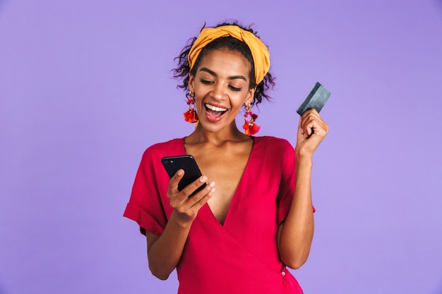 Felice donna africana in abito utilizza lo smartphone mentre si tiene la carta di credito sul muro viola
