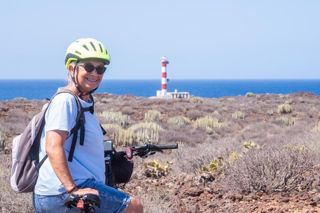 Felice donna anziana attiva dai capelli bianchi in bicicletta fuori strada con bicicletta elettrica in mare anziana donna caucasica in vacanza estiva faro all'orizzonte