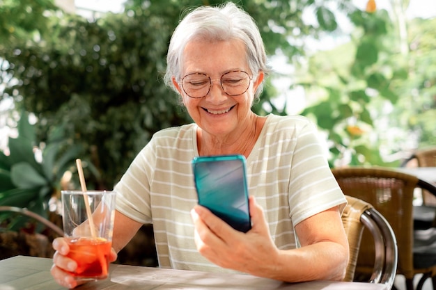 Felice donna anziana attraente seduta all'aperto in un tavolo della caffetteria con in mano un bicchiere e guardando il telefono cellulare signora anziana che si rilassa godendo della tecnologia e del sociale