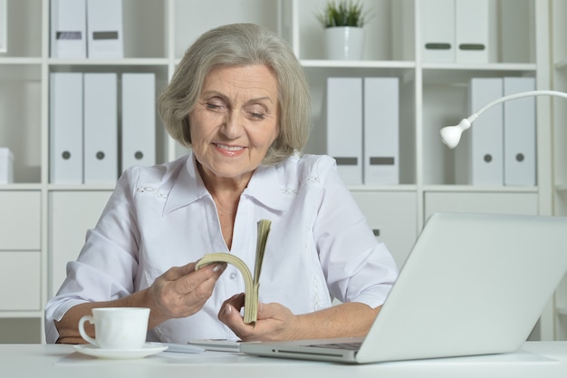 Felice donna anziana che lavora con laptop e soldi in ufficio