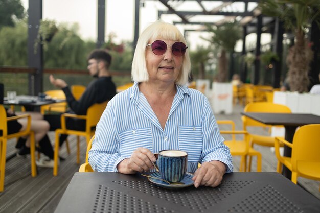 Felice donna anziana con abiti eleganti e occhiali da sole che beve caffè al caffè di strada anziano