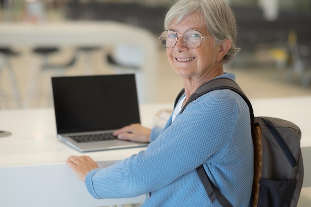 Felice donna anziana in aeroporto in attesa della partenza del volo utilizzando il laptop sulla scrivania bianca Concetto di viaggiatori viaggiatori che tengono zaino