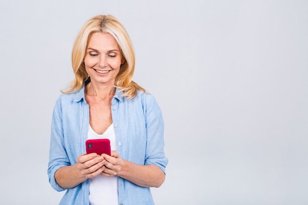 Felice donna anziana matura che tiene in mano lo smartphone utilizzando app online mobili, sorridente vecchia signora di mezza età che scrive messaggi sms in chat sul telefono guardando il cellulare isolato su sfondo grigio bianco