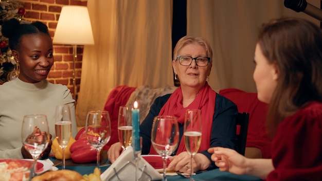 Felice donna anziana seduta al tavolo da pranzo di Natale con i membri della famiglia mentre guarda la fotocamera. Anziano festivo che celebra le vacanze tradizionali invernali con i parenti stretti a casa.