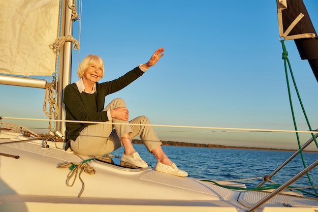 Felice donna anziana seduta sul lato della barca a vela o del ponte di yacht che galleggia nel calmo mare blu