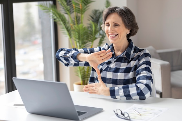 Felice donna anziana sorda usa la lingua dei segni durante la videochiamata utilizzando il laptop mentre è seduta in ufficio a casa