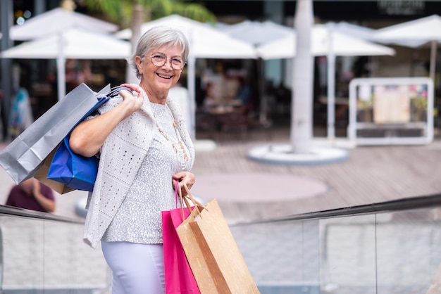 Felice donna anziana sulla scala mobile del centro commerciale che trasporta le borse della spesa guardando la fotocamera sorridendo consumismo vendita cliente concetto di shopping Venerdì nero