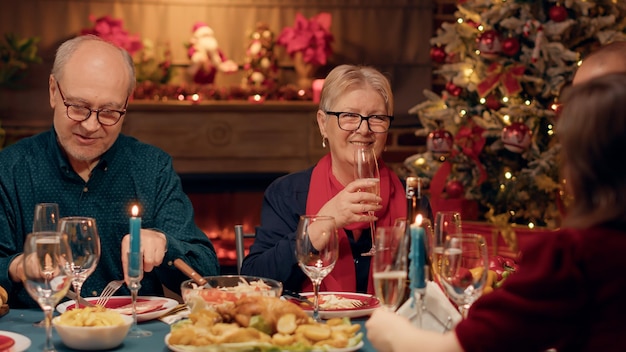 Felice donna anziana tintinnante bicchieri con giovane mentre si gode la cena di Natale in famiglia. Persona anziana sorridente che beve champagne mentre celebra la festa invernale con i parenti.