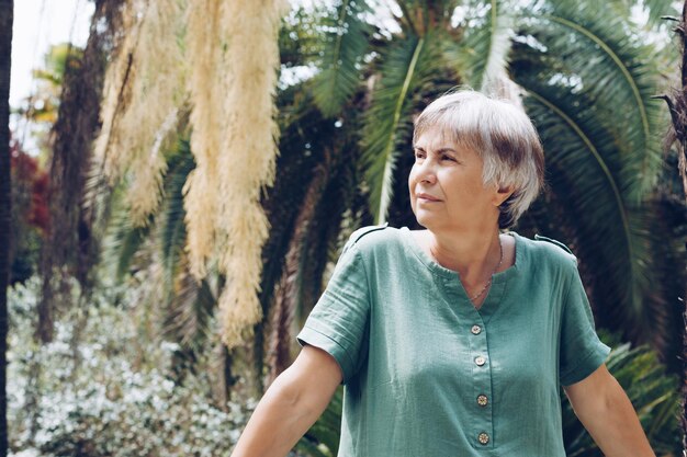 Felice donna anziana vestita con un abito di lino verde si trova di fronte alle palme nel giardino bodanico