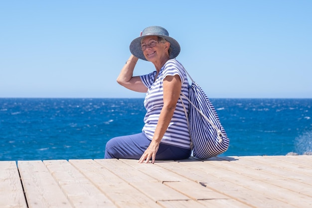 Felice donna anziana vestita di blu seduta al mare tenendo il cappello per timore che voli via Signora matura con occhiali godendo la libertà e rilassarsi in vacanza