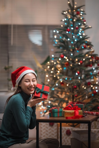 Felice donna asiatica con cappello di babbo natale seduto e mostrando molte scatole regalo con albero di natale a casa.