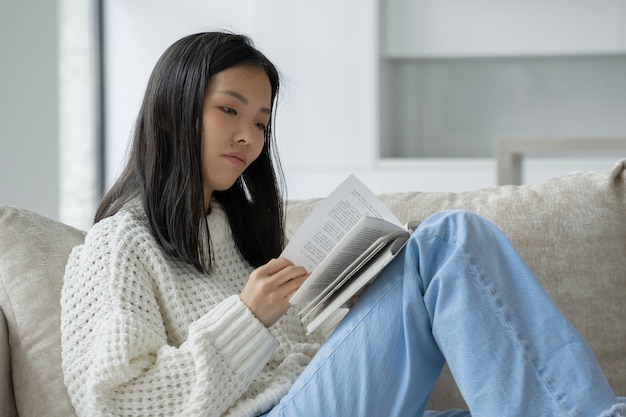 Felice donna asiatica con un maglione bianco seduta sul divano e leggendo un libro a casa