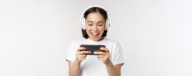 Felice donna asiatica in cuffia guardando smartphone guardando video sul cellulare e sorridente in piedi su sfondo bianco
