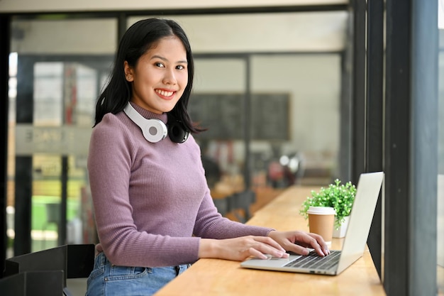 Felice donna asiatica libera professionista si siede a un tavolo contro la finestra nella caffetteria con il suo computer portatile