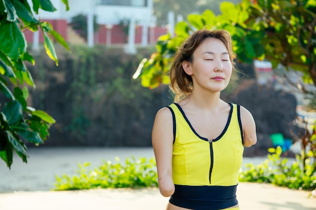 Felice donna asiatica senza braccia che fa meditazione yoga sulla spiaggia tropicale