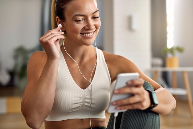 Felice donna atletica che regola gli auricolari mentre si utilizza il telefono cellulare e si prepara per l'allenamento sportivo a casa