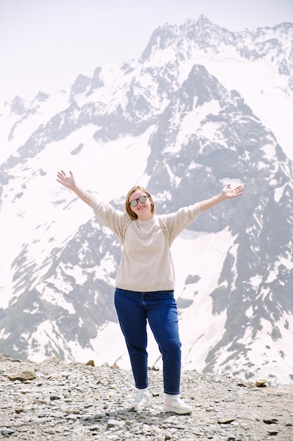 Felice donna attraente in occhiali da sole si erge su una roccia con le mani alzate Felice turista in montagna