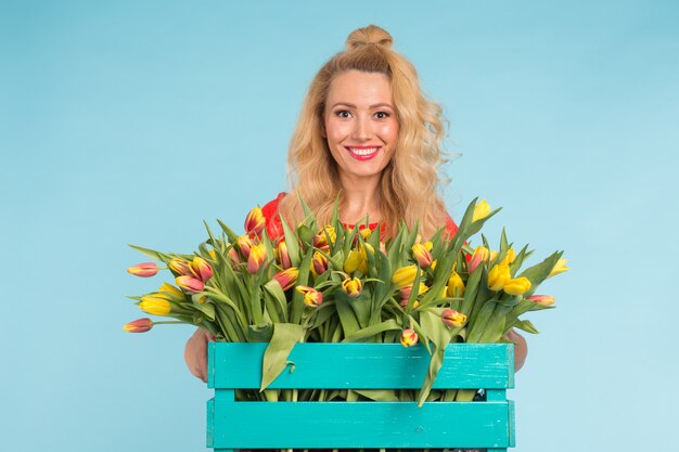 Felice donna bionda caucasica fioraio ridendo e tenendo grande scatola di tulipani su sfondo blu