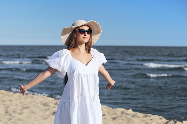Felice donna bionda è sulla spiaggia dell'oceano in abito bianco e occhiali da sole, a braccia aperte.