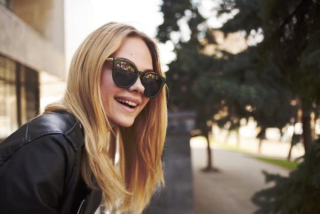 Felice donna bionda in giacca di pelle alla moda occhiali natura parco strada