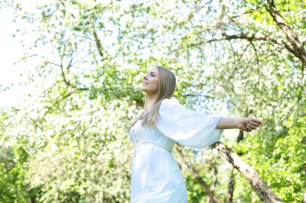 Felice donna bionda in un abito bianco su uno sfondo di un giardino fiorito primaverile con le mani alzate al cielo