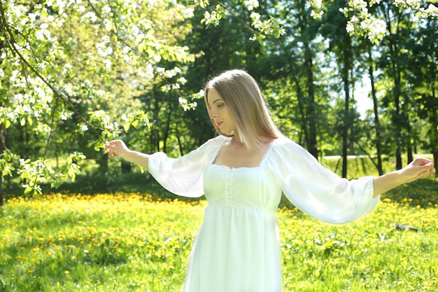 Felice donna bionda in un abito bianco su uno sfondo di un giardino fiorito primaverile con le mani alzate al cielo