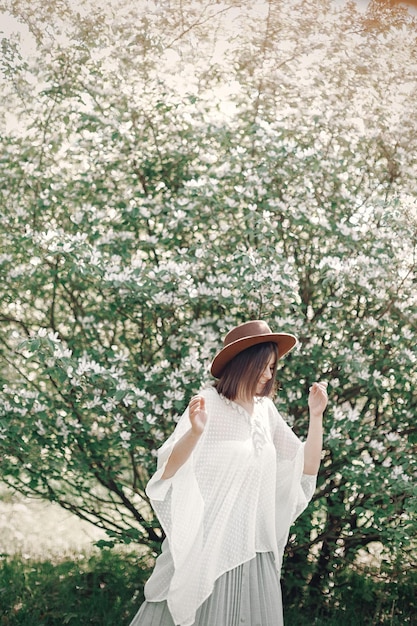Felice donna boho in cappello che si diverte alla luce del sole vicino all'albero bianco in fiore nel parco primaverile Elegante ragazza hipster che si gode la primavera e balla Momento atmosferico di felicità