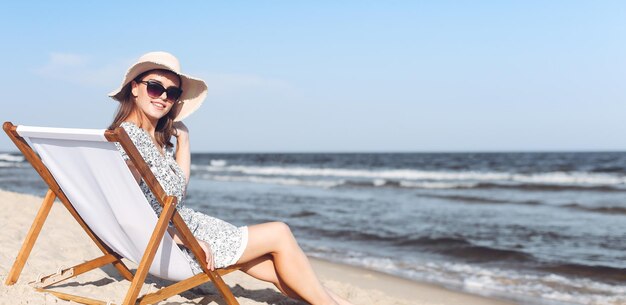 Felice donna bruna che indossa occhiali da sole e cappello che si rilassa su una poltrona di legno sulla spiaggia dell'oceano