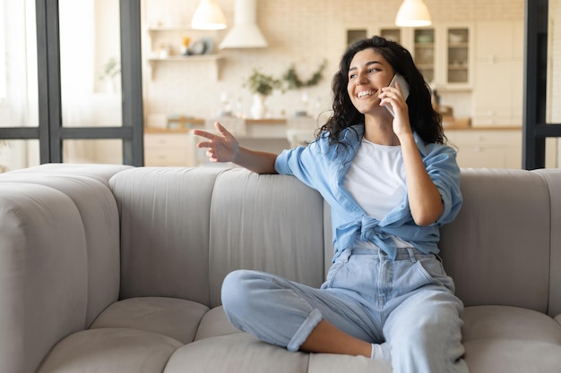 Felice donna bruna millenaria seduta sul divano che effettua chiamate sul telefono cellulare a casa spazio libero