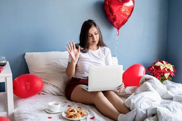 felice donna bruna seduta nel letto con palloncini a forma di cuore rosso lavorando sul portatile da casa