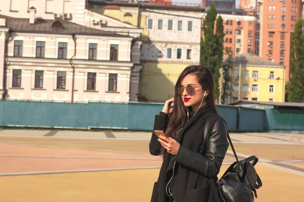 Felice donna bruna turista ascolta musica sul telefono cellulare, in piedi per strada con lo zaino