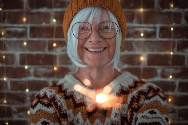 Felice donna caucasica anziana con occhiali e maglione invernale che tiene la luce scintillante anziana signora gioiosa che celebra il Natale e il nuovo anno scintillante nella notte