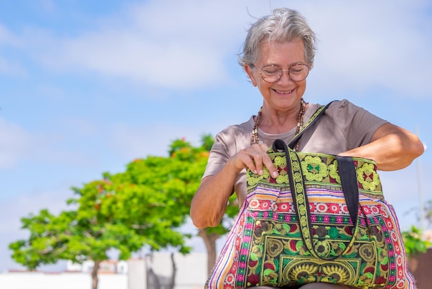 Felice donna caucasica senior seduta nel parco guardando nella sua borsa fatta a mano sorridente anziana donna dai capelli grigi che si rilassa all'aperto in una giornata estiva