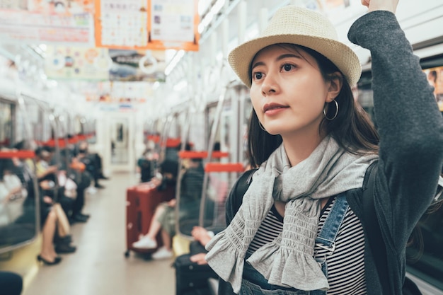 Felice donna cinese in una metropolitana. Elegante viaggiatore femminile con cappello guarda da parte in piedi nella tranquilla maniglia della tenuta della mano del treno della metropolitana. bella signora sorridente che vede fuori dalla finestra con la gente del posto seduta sul sedile