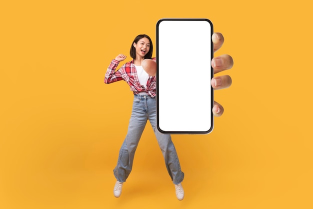 Felice donna coreana che mostra lo schermo vuoto dello smartphone mentre salta sopra il mockup del collage di sfondo giallo dello studio